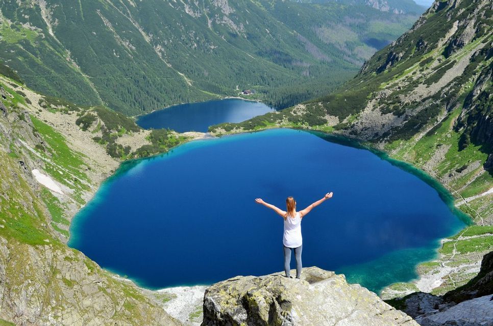A woman stands on a rocky outcrop with arms raised, looking out over two vibrant blue lakes nestled in a green mountain range.