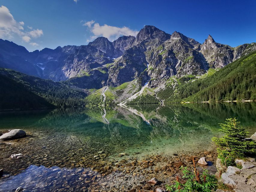 A range of jagged mountains with green slopes reflecting in the still, clear water of an alpine lake.
