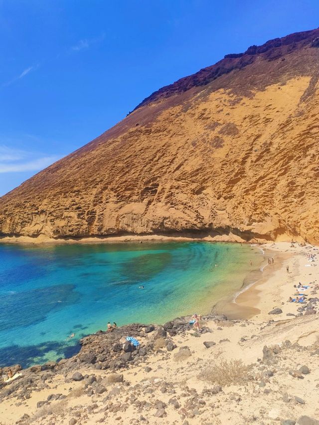 Una cala de playa arenosa con aguas cristalinas de color turquesa al pie de un acantilado empinado de color ocre bajo un cielo azul brillante.