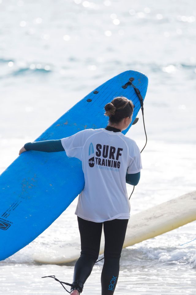 Una mujer en traje de neopreno, vista de espaldas, sostiene una tabla de surf azul grande mientras está de pie en el agua poco profunda del océano.