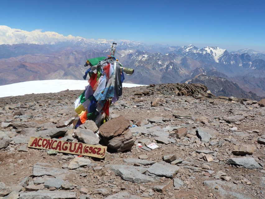 A wooden sign for Aconcagua rests on a rocky summit next to a post with colorful flags, with snowy mountain peaks in the distance.