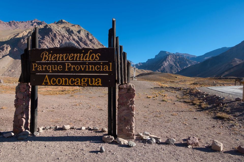 A wooden sign for 'Parque Provincial Aconcagua' at the entrance to a mountainous, arid landscape under a clear sky.
