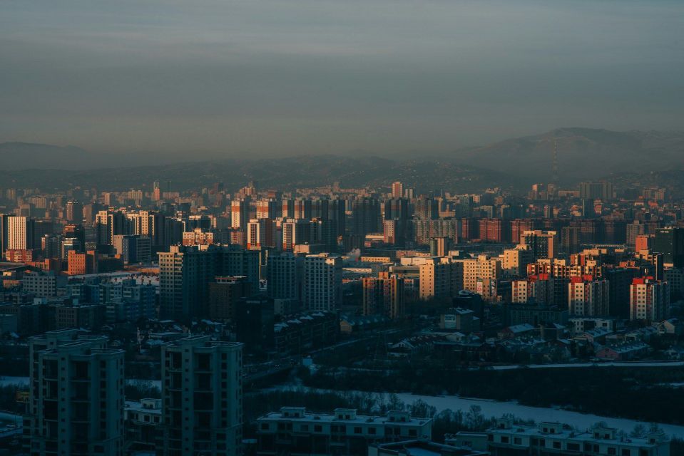A dense cityscape with numerous buildings illuminated by warm, low sunlight, with hazy mountains in the background.