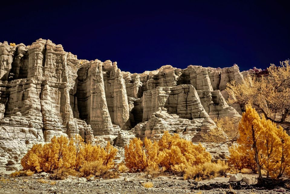 Jagged, light-colored rock formations rise above yellow trees on a rocky terrain under a deep blue sky.