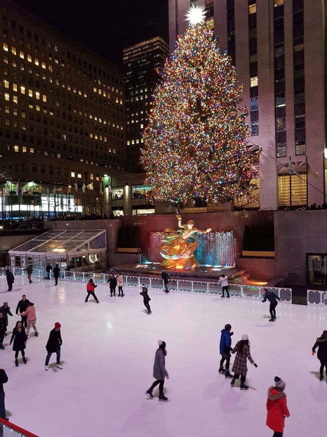 Personas patinando sobre hielo de noche en una pista al aire libre frente a un gran árbol de Navidad iluminado y altos edificios de la ciudad.