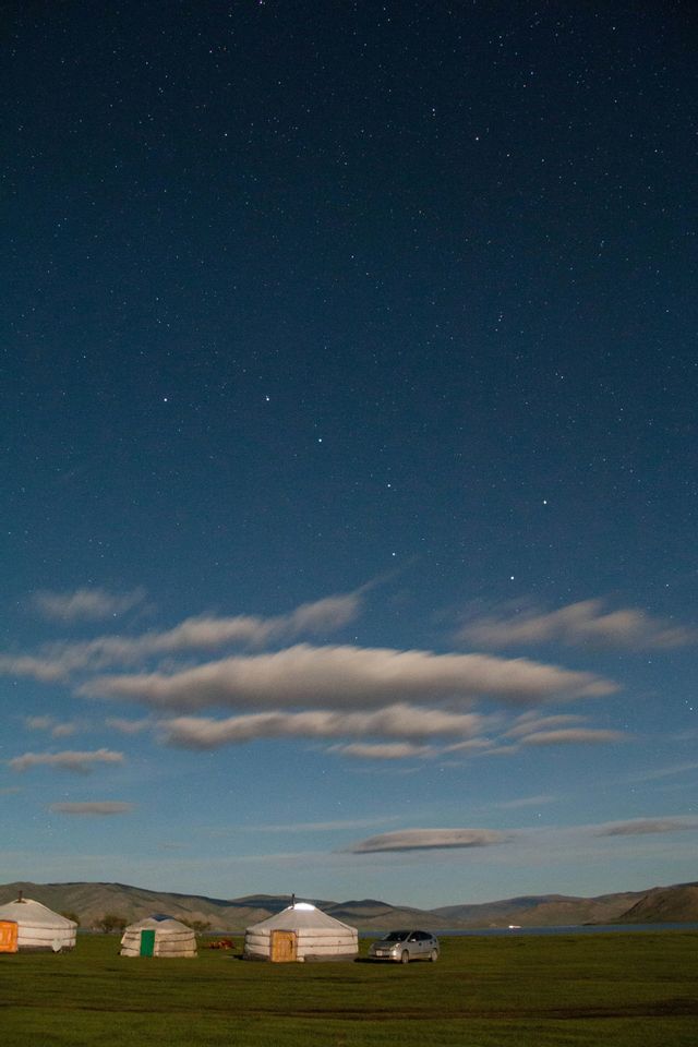 Einige Jurten und ein Auto stehen auf einem grünen Feld unter einem weiten, sternenklaren Nachthimmel mit langen, weißen Wolken.