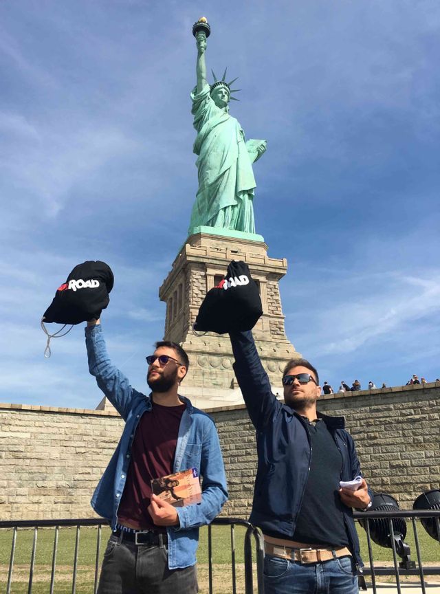 Deux hommes d'un voyage de groupe WeRoad brandissant des sacs noirs devant la Statue de la Liberté sous un ciel bleu.