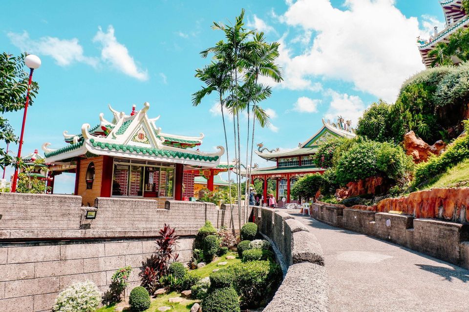 Templi sontuosi con tetti di tegole verdi circondati da giardini lussureggianti, palme e sentieri in pietra sotto un cielo blu.