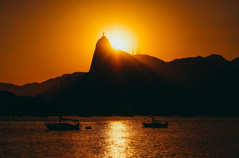 El sol se pone detrás de una montaña coronada con una estatua, silueteando la cima y proyectando un brillo naranja sobre los barcos en una bahía.