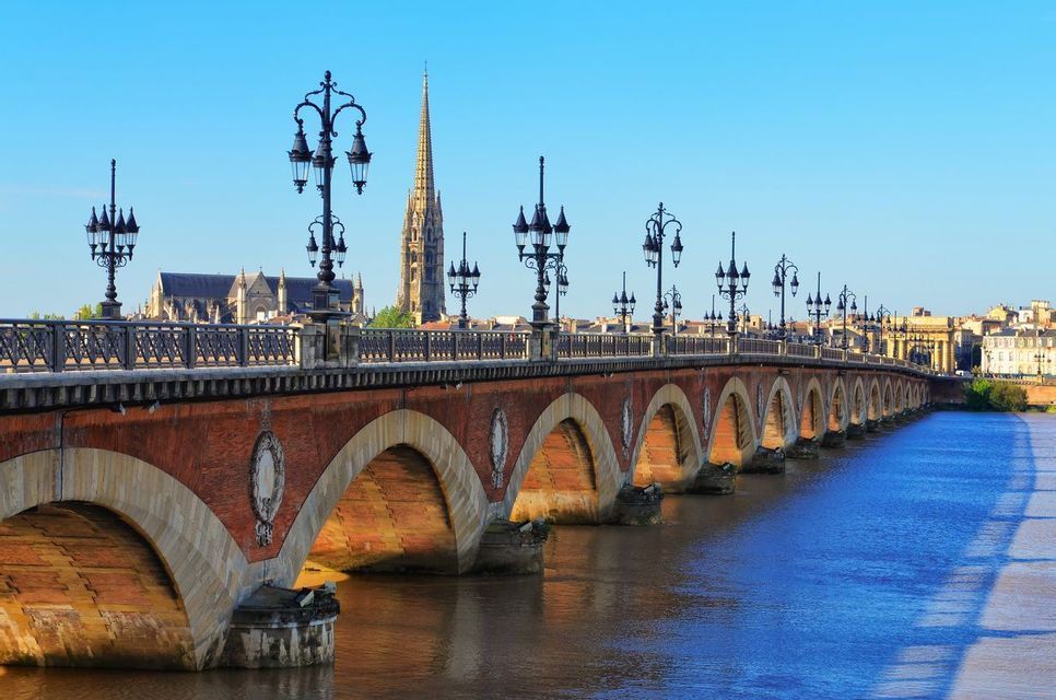 Un ponte ad arco in pietra con lampioni decorativi attraversa un ampio fiume, con lo skyline di una città visibile in lontananza.