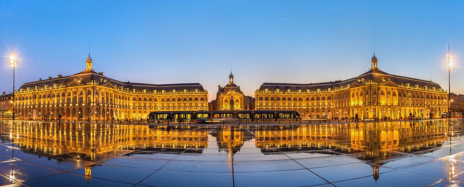 Una vista panoramica di un edificio storico illuminato che si riflette in una piazza bagnata al tramonto, con un tram in movimento.
