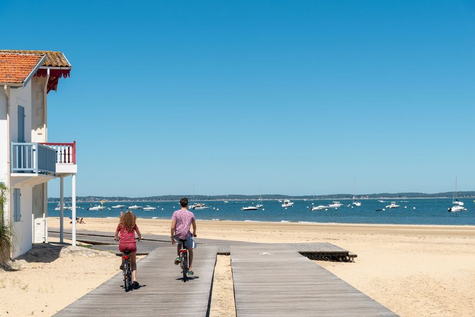 Un uomo e una donna pedalano su una passerella di legno lungo una spiaggia sabbiosa, con numerose barche sull'acqua.