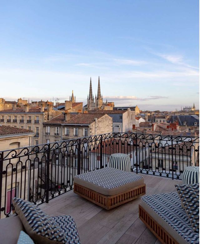 Una terrazza panoramica arredata con ringhiera in ferro battuto si affaccia su un paesaggio urbano storico con guglie di chiesa sotto un cielo sereno.
