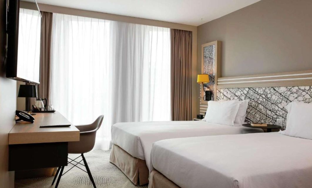 Una moderna camera d'hotel con due letti singoli con lenzuola bianche, una scrivania e una grande finestra con tende trasparenti.