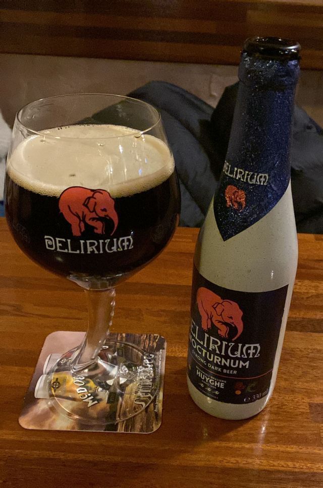 Un bicchiere di birra scura Delirium Nocturnum con una schiuma densa, posto accanto alla sua bottiglia abbinata su un tavolo di legno.