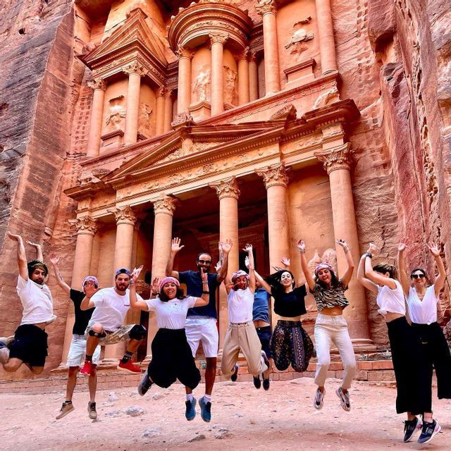 Un voyage de groupe WeRoad, sautant pour une photo devant un grand temple antique sculpté dans une falaise de roche rouge.