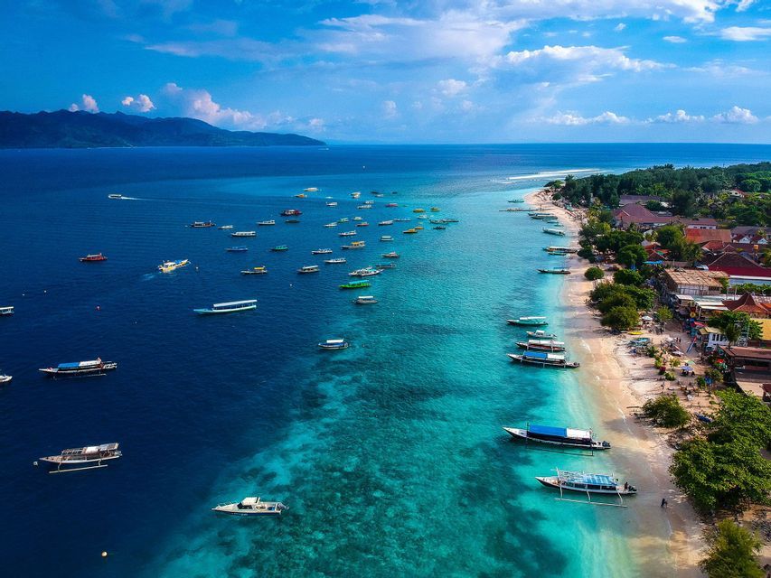 Una vista aérea de muchos barcos flotando en agua turquesa a lo largo de una playa de arena, con montañas en el horizonte bajo un cielo parcialmente nublado.