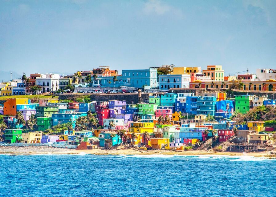Un gruppo di edifici dai colori vivaci su una collina che domina una spiaggia sabbiosa e l'oceano blu sotto un cielo limpido