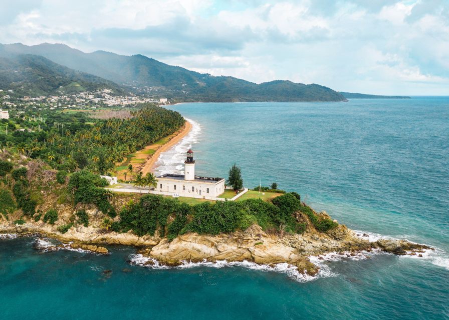 Vista aerea di un faro bianco su un promontorio verde e roccioso accanto a una spiaggia sabbiosa costeggiata da palme, con montagne sullo sfondo.
