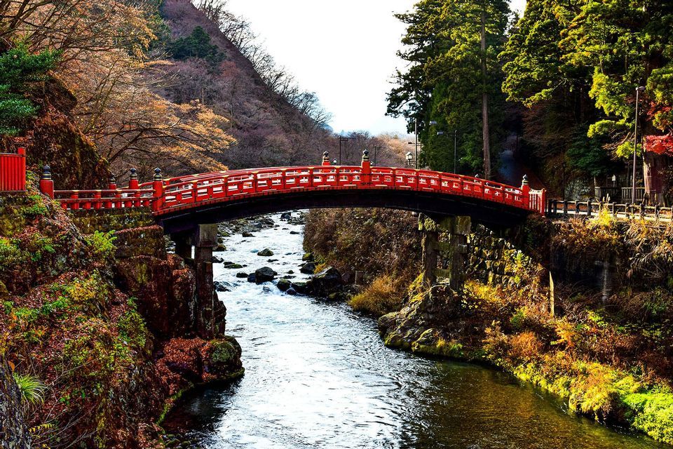 Un puente rojo y ornamentado con arcos cruza un río rocoso que fluye por un valle boscoso con follaje de otoño.