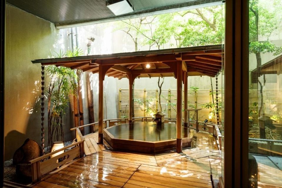 Un onsen tradicional de madera en un patio exterior, con una glorieta, tarima mojada y plantas de bambú.