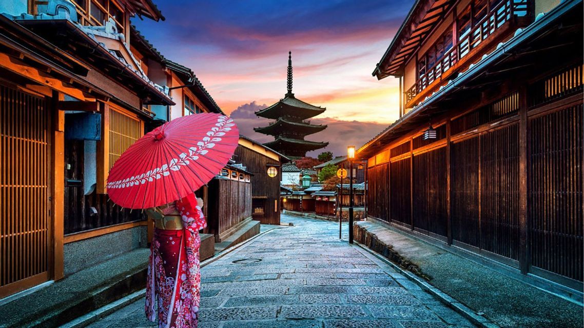 Una persona con un kimono sostiene un paraguas rojo en una calle empedrada bordeada de casas tradicionales y una pagoda al atardecer.