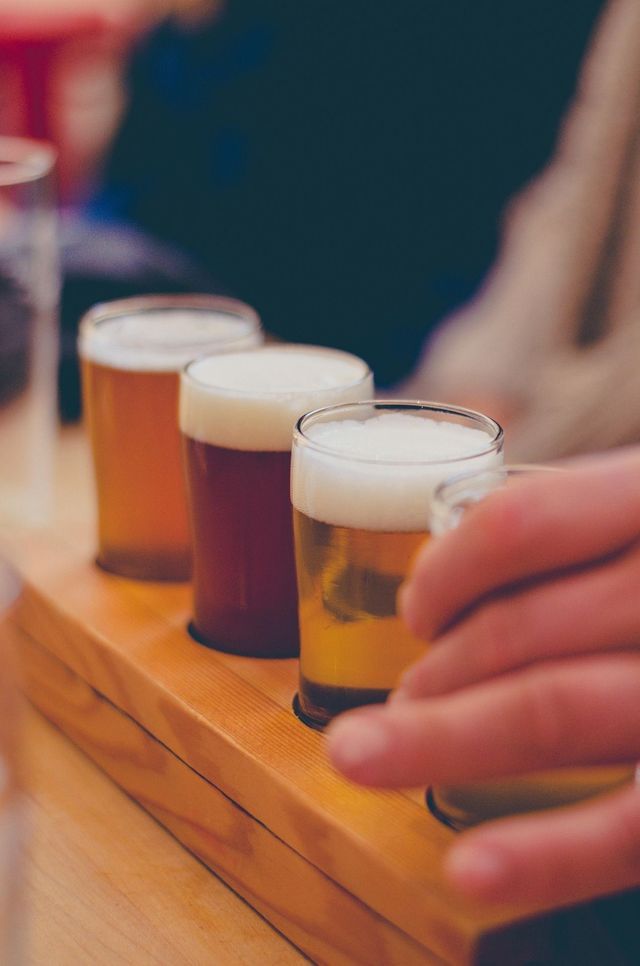 Primer plano de una cata de cervezas con cuatro vasos de diferentes cervezas en un soporte de madera, mientras una mano alcanza uno.