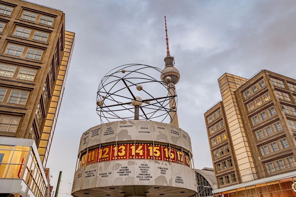 Una toma en ángulo bajo de un gran reloj mundial y una alta torre de televisión, enmarcada por edificios urbanos bajo un cielo nublado.