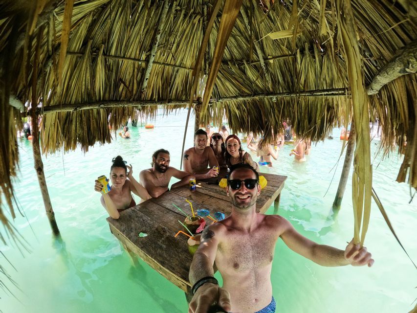 Un groupe WeRoad en voyage prend un selfie en buvant à une table partiellement immergée dans une eau turquoise sous un toit de chaume.