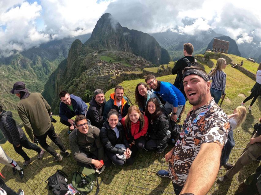 Un groupe WeRoad prend un selfie souriant sur un point de vue herbeux, surplombant des ruines antiques nichées dans des montagnes escarpées et verdoyantes.