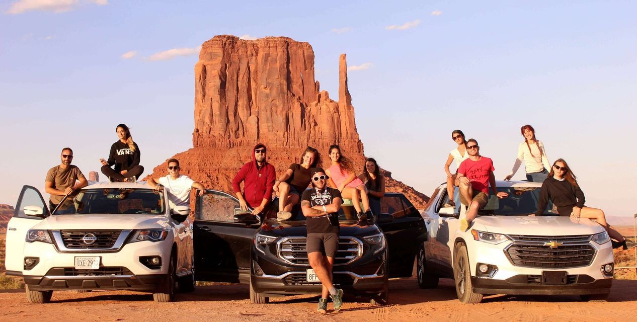 A WeRoad group trip posing on and around three cars in a desert landscape with red rock formations in the background.