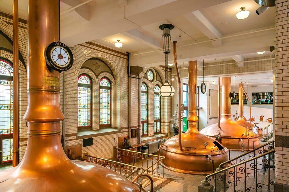 El interior de una cervecería tradicional con grandes calderos de cobre para la elaboración, paredes de azulejos blancos y ventanas arqueadas de vidrieras.