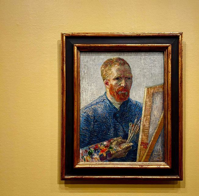 Ein gerahmtes Gemälde eines Selbstporträts von Vincent van Gogh, eine Palette und Pinsel haltend, hängt an einer gelben Wand.