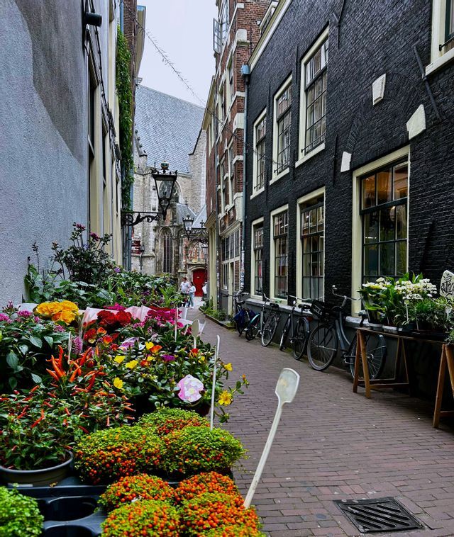 Flores y plantas de colores a la venta en un estrecho callejón empedrado, bordeado por edificios históricos y bicicletas estacionadas.