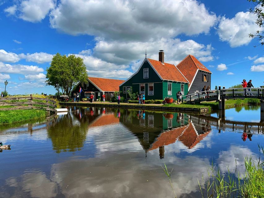 Des maisons en bois vertes aux toits de tuiles orange se reflètent dans l'eau calme d'un canal sous un ciel bleu nuageux.