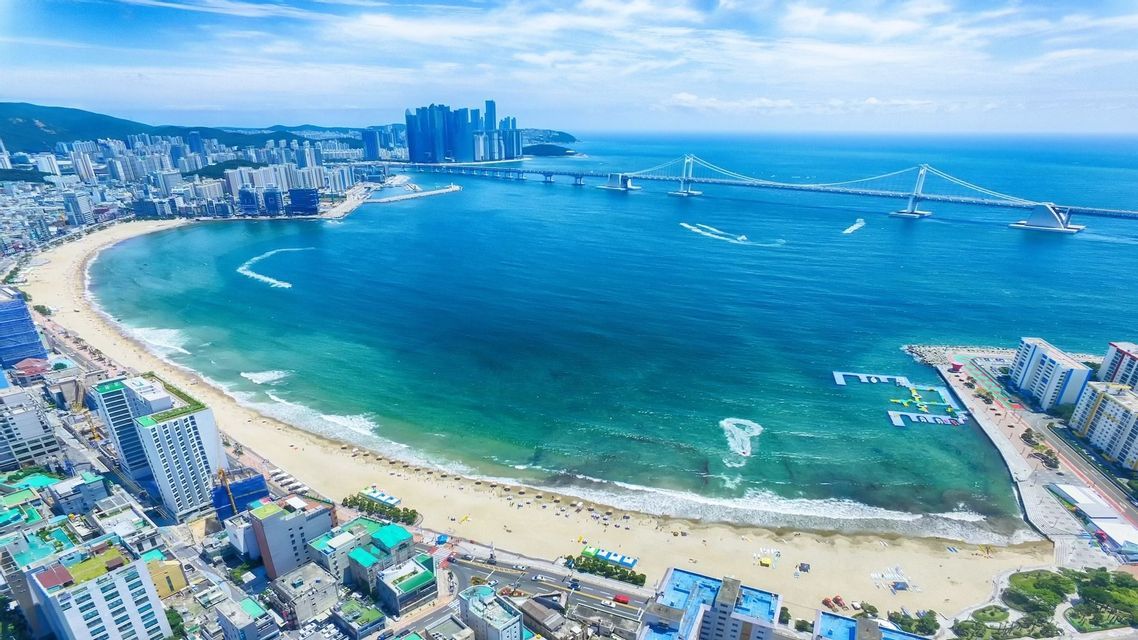 Vista aerea di una vivace città costiera con una spiaggia sabbiosa che si curva attorno a una baia, con un lungo ponte in lontananza.