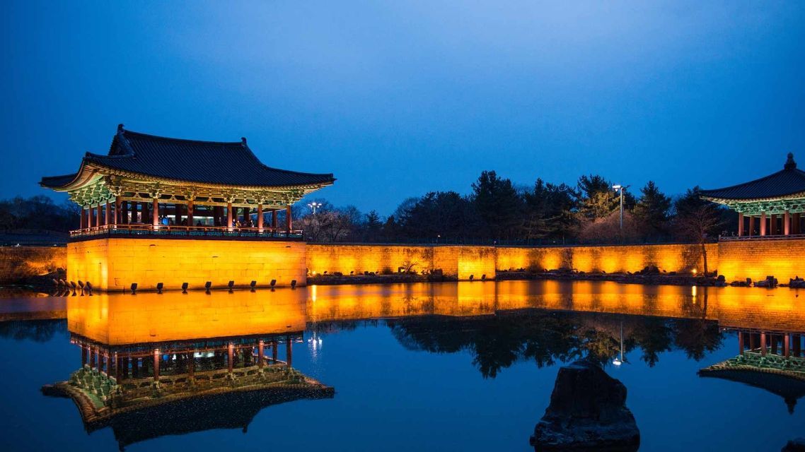 Un tradizionale padiglione e muro dell'Asia orientale, illuminato da luci gialle, che si riflette su un laghetto calmo al crepuscolo.