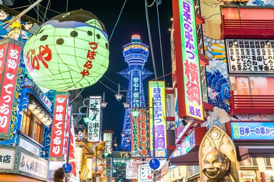 Una vivace strada giapponese di notte, illuminata da insegne al neon colorate, una grande lanterna a forma di pesce palla e una torre illuminata in lontananza.