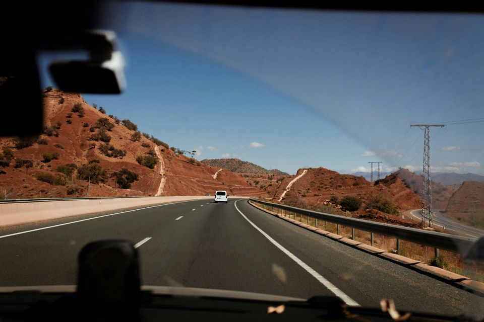 Vista dall'interno di un'auto su un'autostrada che si snoda tra colline aride e rossastre, con un furgone bianco in lontananza.