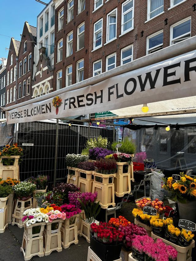 Un puesto de mercado de flores al aire libre con un cartel de 'Flores Frescas', mostrando ramos coloridos en cajas en una calle de la ciudad.