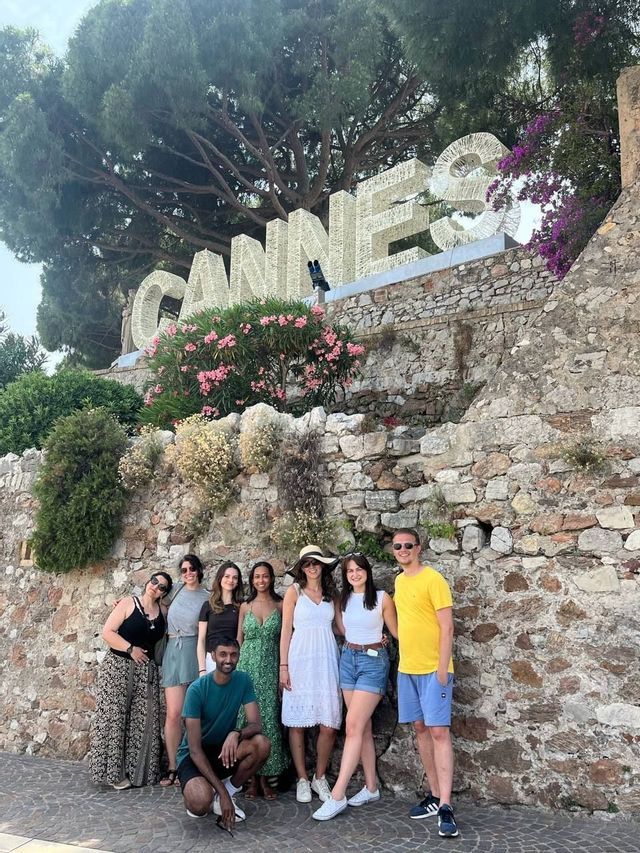 A WeRoad group trip poses for a photo in front of a large 'CANNES' sign on a stone wall with pink flowers.