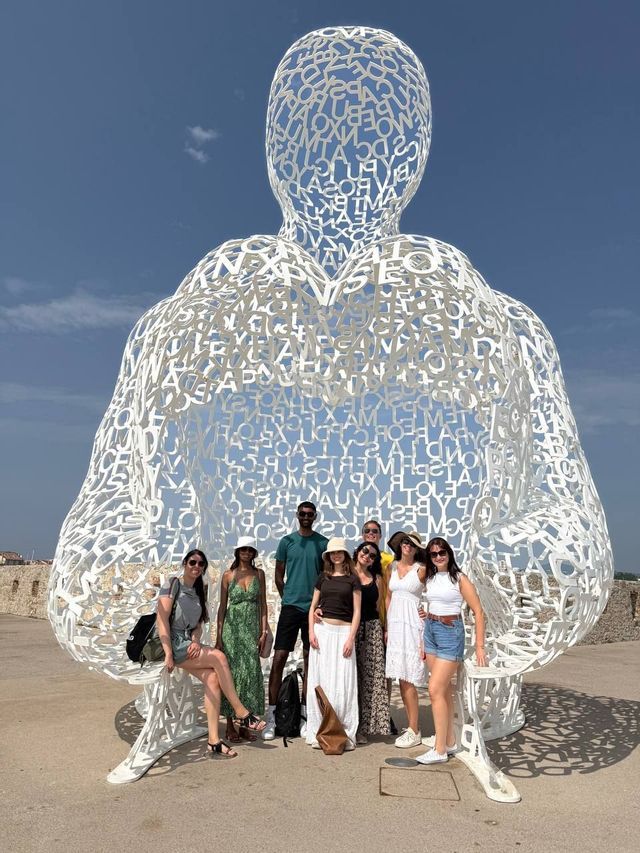 A WeRoad group trip poses for a photo in front of a large, white, human-shaped sculpture made of letters under a clear blue sky.