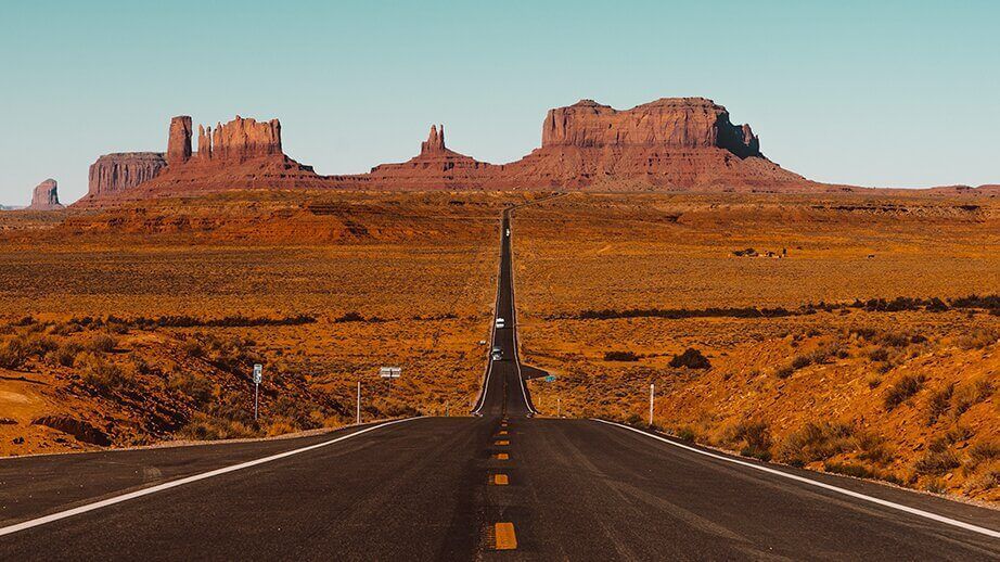 A long, straight road stretches through a desert landscape towards towering red rock buttes under a clear blue sky.