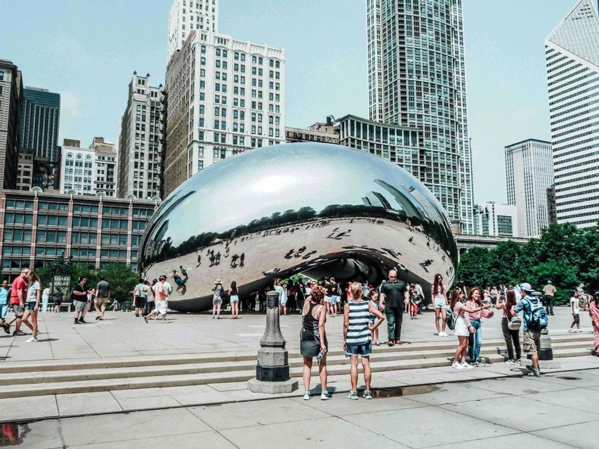A crowd of people gathers around a large, reflective, bean-shaped sculpture in a city plaza, with skyscrapers in the background.