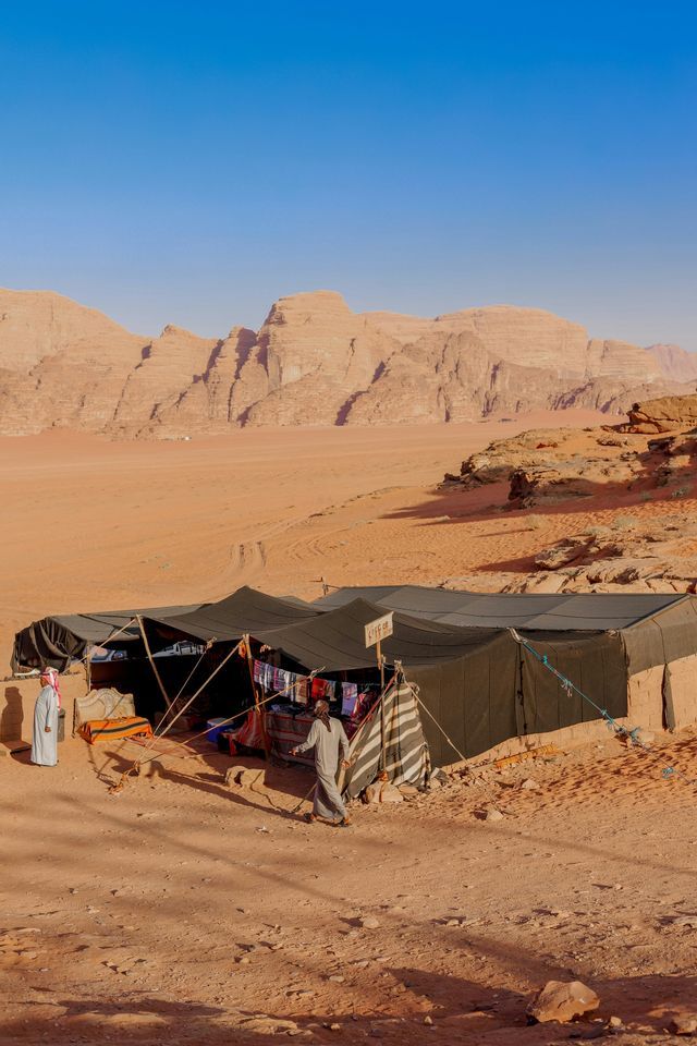 Due persone in abiti tradizionali fuori da una bancarella tendata in un deserto con grandi formazioni rocciose.
