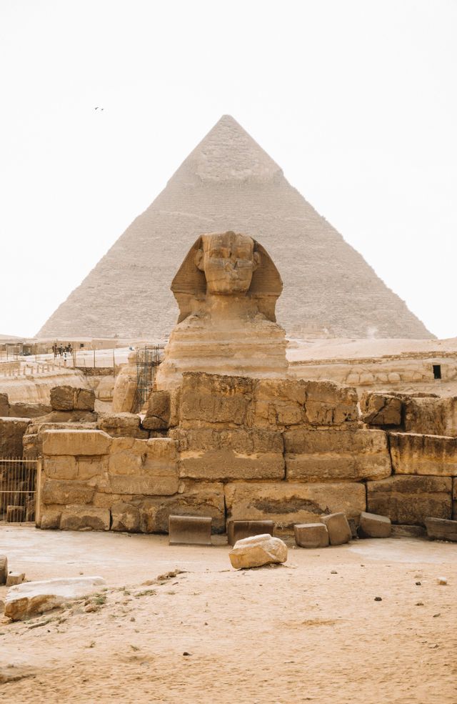 Un'imponente statua antica di sfinge si erge di fronte a una massiccia piramide di pietra, circondata da rovine su terreno sabbioso.