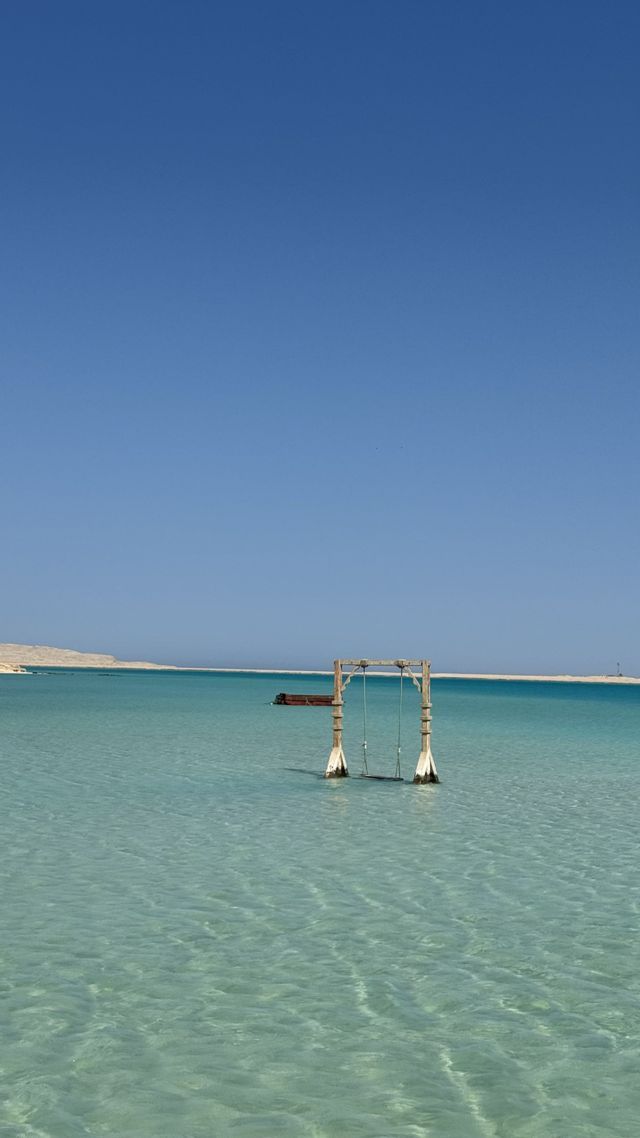 Un'altalena di legno si erge in acque poco profonde e limpide di colore turchese sotto un cielo di un azzurro intenso, con una costa sabbiosa in lontananza.
