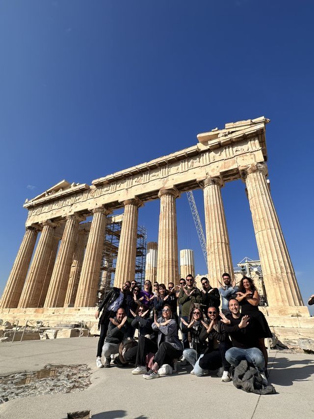 Un viaggio di gruppo WeRoad in posa per una foto davanti a un antico tempio con grandi colonne sotto un cielo azzurro chiaro.