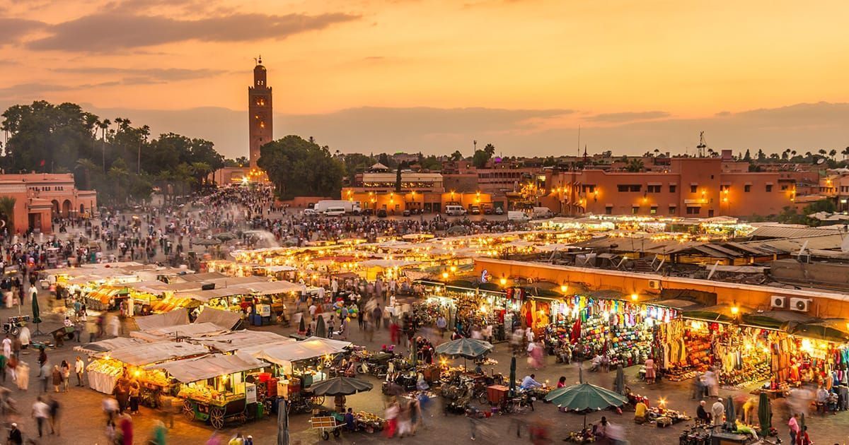 Vista dall'alto di una vivace piazza del mercato cittadino al tramonto, gremita di folla e bancarelle luminose, con una torre sullo sfondo.