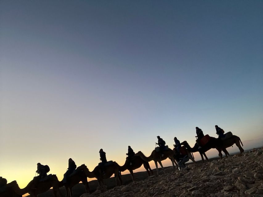 Sagome di un gruppo WeRoad in viaggio che cavalca cammelli in fila attraverso un paesaggio roccioso al tramonto.